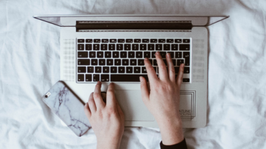 hands typing on a laptop on top of a duvet