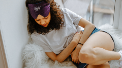 Young woman in pyjamas curled up by a window holding her stomach due to period cramps